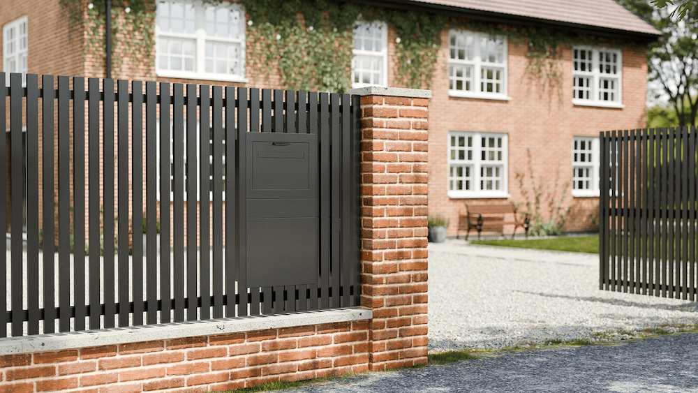 Modern black post and parcel box mounted on a black slatted fence in front of a brick house with white-framed windows.