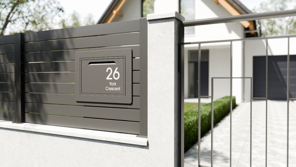 Anthracite fence-mounted parcel box installed on a modern slatted fence beside a driveway entrance.