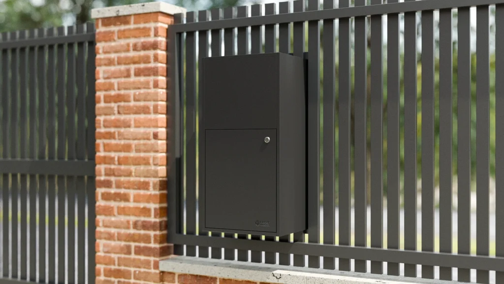 Black fence-mounted parcel box securely installed on a modern metal fence beside a brick pillar.